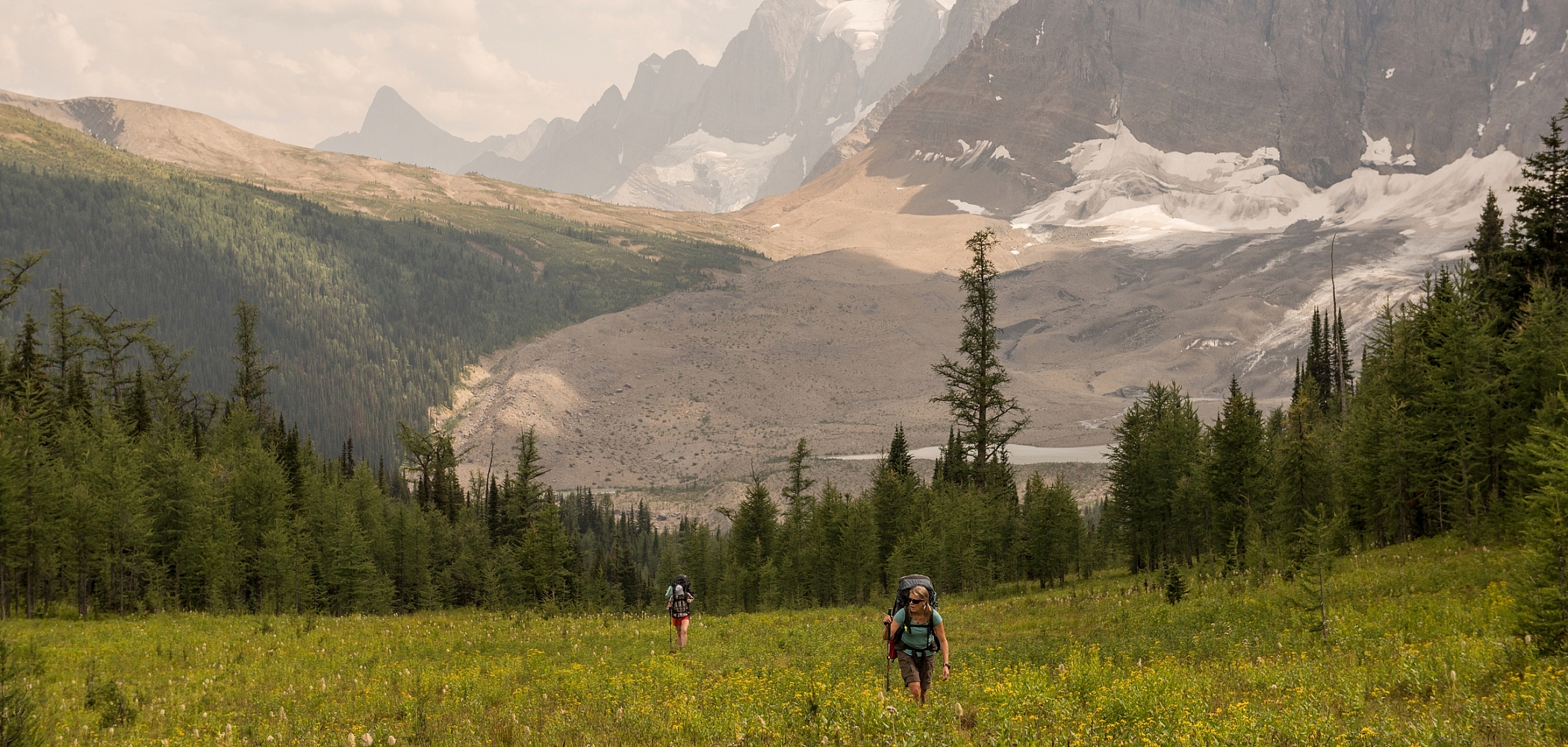 Two people with big backpacks walk toward the camera through a grassy field. Behind them is a ring of evergreen trees, and beyond those are rugged mountain peaks and glaciers.
